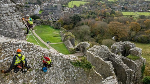 Stone masons on high ropes, walking along the top of the ruins with Corfe Castle village in the background